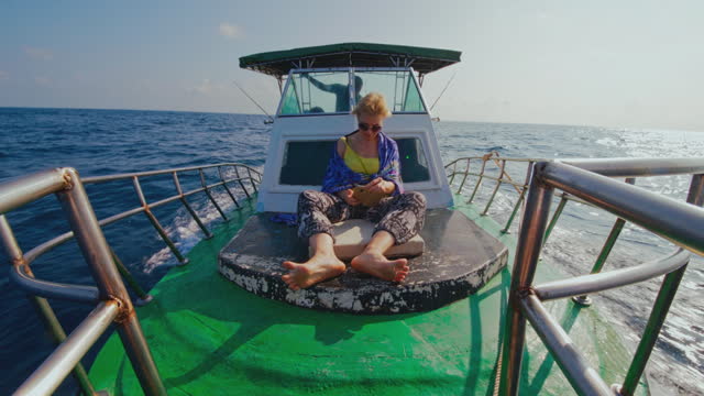 A 50-years-old active European woman, a tourist, is resting and checking her smartphone on the deck of a small fishing boat during the deep sea fishing trip in Sri Lanka when a captain is ruling the ship from a deckhouse in the backdrop.
