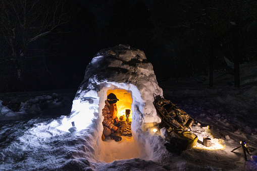 A lone mid age woman inside an igloo heating up water and warming up by a gas heater at night in a snowy winter forest.