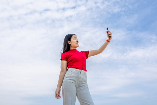 Image of low angle view,Relaxing young woman with long hair enjoying beautiful air of nature and taking a photo on mobile with smile happily, Girl posing for photo outdoor.