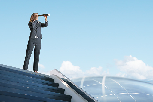 A businesswoman at the top of a staircase peering through a spyglass at a globe in the distance. Standing atop a futuristic staircase, she scopes out the future contemplating innovation. A soft halo of light emanates off the wistful globe, with a grid pattern traversing the atmosphere.