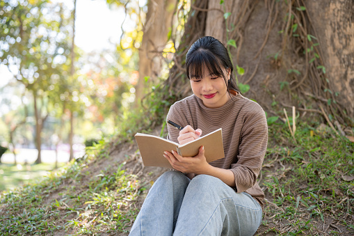 A positive and pretty Asian woman in casual clothes is keeping her diary or jotting down her ideas in a book while resting, sitting under a tree in a park. people and leisure concepts