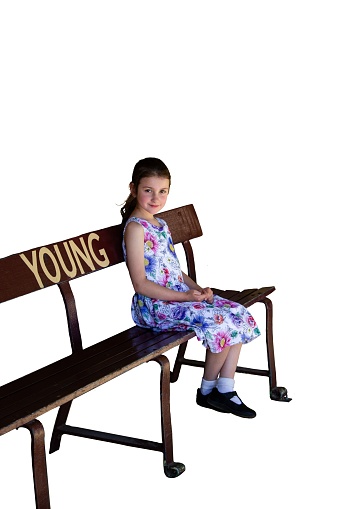 Young girl sitting on a bench, 6 year old girl in floral dress, smiling, white background, copy space, bench with Young written on it,