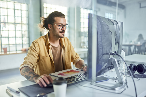 Smiling male graphic designer working on PC in the office. The view is through glass.