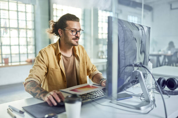 Young male programmer working on desktop PC in the office. Smiling male graphic designer working on PC in the office. The view is through glass. graphic designer stock pictures, royalty-free photos & images
