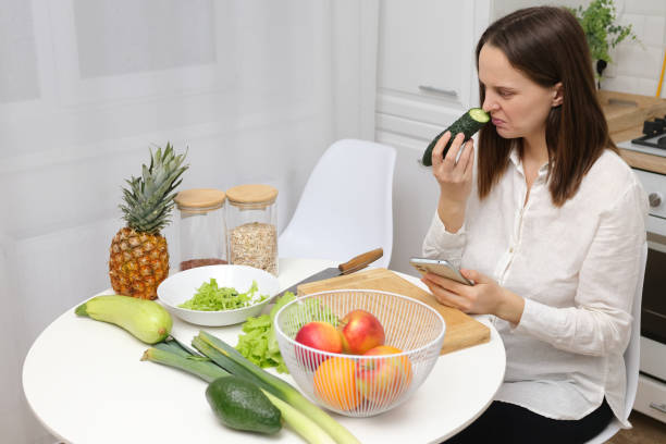 istock Confused puzzled Caucasian brown haired woman sitting at kitchen table with fruit and vegetables smelling not fresh cucumber from refrigerator or freezer 1976936324