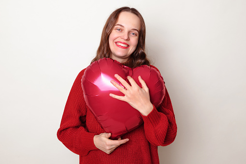 Cute smiling pretty brown haired adult woman wearing red sweater embracing heart shape balloon posing isolated over gray background celebrating St. Valentines Day