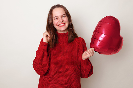 Attractive romantic brown haired adult woman wearing red sweater holding heart shape balloon posing isolated over gray background celebrating Valentines day flirting looks shy