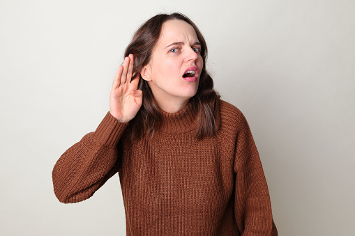 Caucasian woman wearing brown jumper standing isolated over gray background holding hand near ear and listening carefully having hearing problems deafness in communication