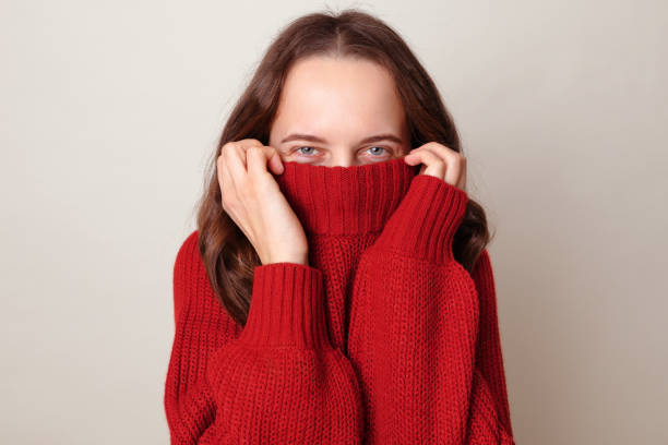 istock Brown haired woman pulling her red trendy sweater over head having fun being childish disappearing in her clothes looking from underneath with cheerful shy eyes isolated over gray background 1976938475