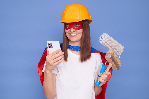 Happy woman painter wearing superhero costume and protective helmet holding painting roller and brush isolated over blue background using smartphone scrolling online checking mobile app