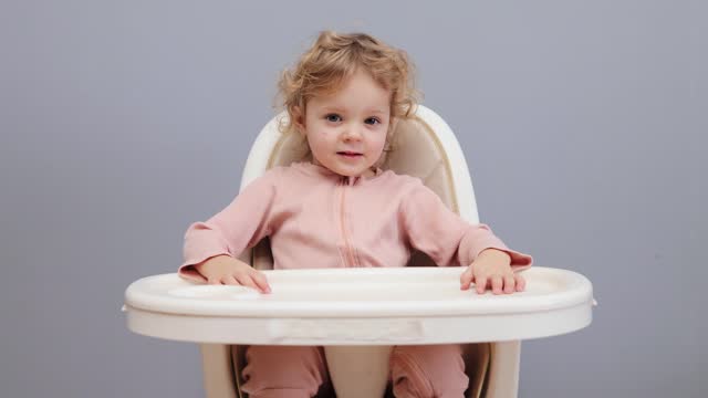 Cheerful blond wavy haired toddler satisfied joyful little girl sitting in highchair waiting her breakfast or dinner isolated over gray background