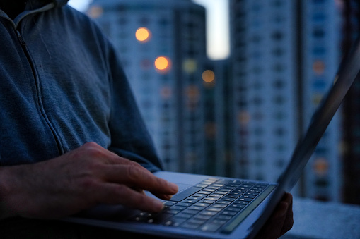 View of a laptop on a balcony with a person’s hands on the keyboard and the city night lights behind.