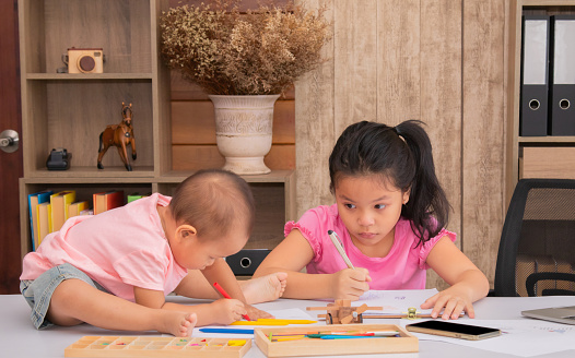 Adorable 2 kids sitting on desk full of crayon colors and white papers enjoy playing together at home, 2 sibling happy drawing with their imagination, elder sister play with naughty younger sister