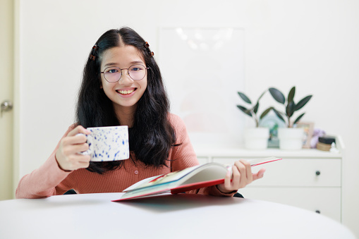 A young, happy Asian girl is depicted sitting at her desk, holding a white mug, and looking at the camera while reading a book. This scene conveys a sense of contentment and enjoyment in the act of reading, combined with the comfort of holding a warm mug.