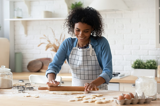 Happy millennial African American woman in apron cooking bakery food in home kitchen, rolling dough, smiling. Blogger, baker, chef enjoying job, making cookies, preparing pastry dessert, biscuit, cake