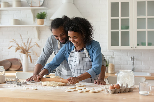 Happy young African couple wearing aprons cooking homemade bakery foods together, kneading, rolling dough for cookies at flour table, talking, laughing. Husband helping wife in home kitchen