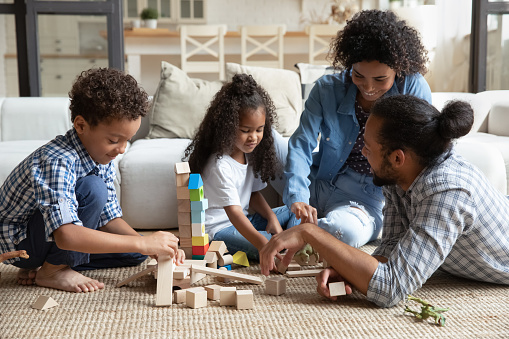 Happy millennial African family couple and little sibling kids constructing toy castle on heating floor at home, building towers from small wooden blocks, playing together, enjoying leisure, playtime