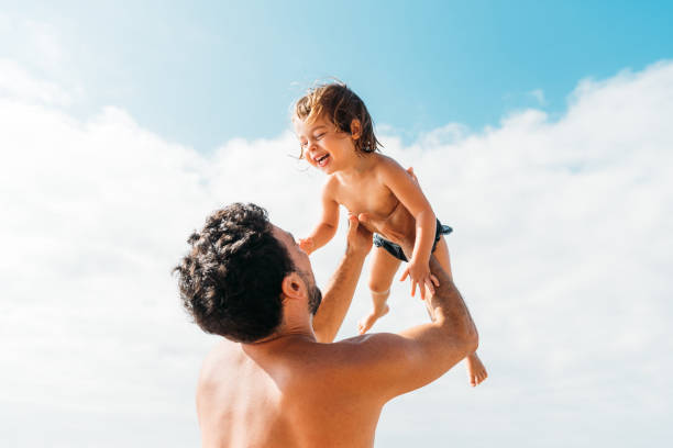 Happy young father holding little kid against cloudy sky Back view of happy shirtless young father lifting little kid while standing against cloudy sky on sunny day half dressed girls pictures stock pictures, royalty-free photos & images
