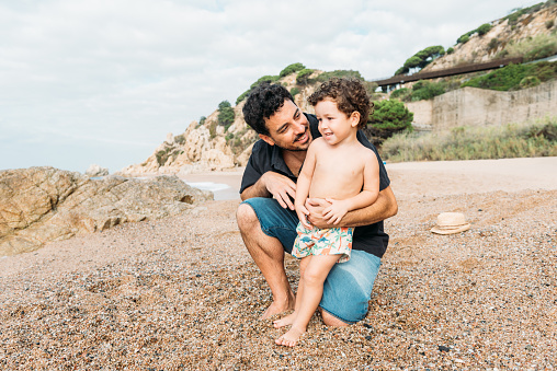 Young smiling father with little kid sitting on sandy beach and hugging cute son while enjoying sunny day against mountains
