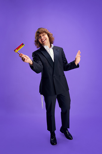 Joyful man in suit holding grogger symbol used to drown out name of Haman villainous character against purple backdrop. Celebration of Purim, festival, holiday, Judaism, religion concept. Ad