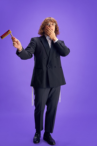 Full length portrait of young scared Jewish man holding grogger symbol used to drown out name of Haman villainous character. Purim, business, festival, holiday, celebration, religion concept.