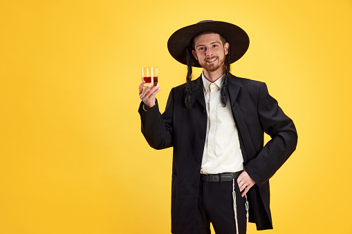 Portrait of smiling cheerful man, Jewish holds glass with alcoholic drink against sunny yellow background. Purim, business, festival, holiday, celebration Pesach or Passover, Judaism, religion concept