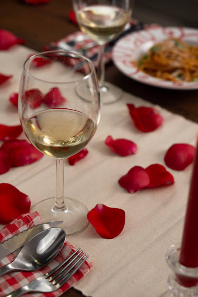 istock Dinner table set for two, with white wine, bolognese spaghetti and red roses' petals on the table. There is a lighted red candle. Vertical image 1977223103