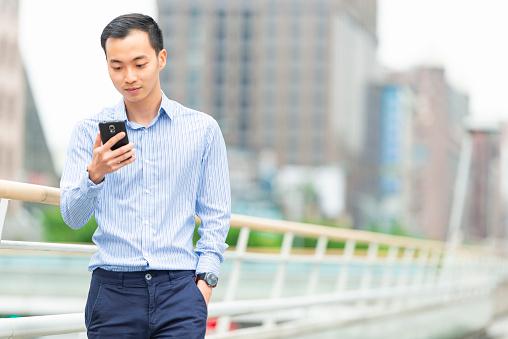 Horizontal color image of a real life Taiwanese white collar professional walking on a footbridge with his phone in Xinyi.