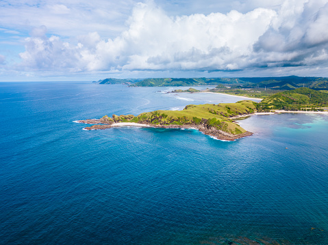 A green coastal island surrounded by deep blue ocean waters. The island is lush and verdant, with a variety of trees and vegetation visible. The ocean is a deep blue, with white waves visible where it meets the island. The sky above is partly cloudy, with white clouds scattered across the bright blue backdrop. The viewpoint is from the ocean, looking towards the island. Shot taken near Lombok island.