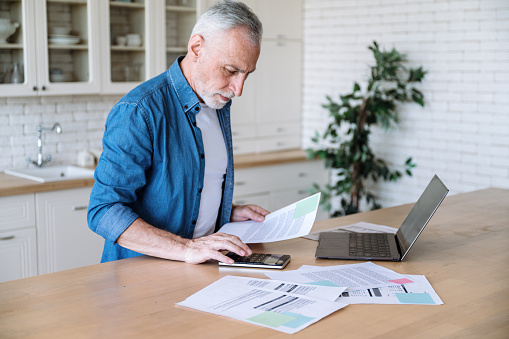 Focused middle aged man making calculations of utility payment holding paper bill invoice in hand at table. Serious businessman doing paperwork for paying taxes online using laptop at home