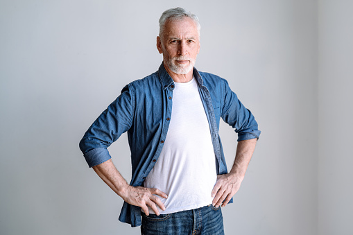 Confident middle aged man wearing denim shirt standing on white background, keeping hands on waist and looking at camera. Portrait of serious mature male entrepreneur in casual clothes