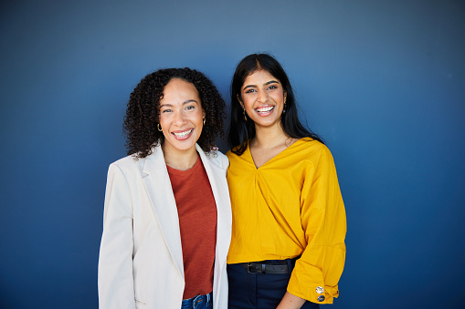 Portrait of two diverse young businesswomen laughing together while standing in front of a blue wall in a modern office