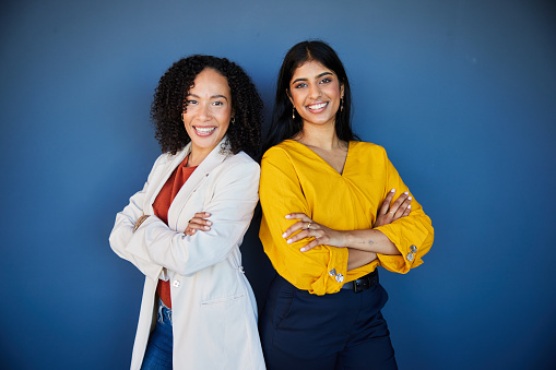 Portrait of two diverse young businesswomen smiling and standing confidently with their arms crossed in front of a blue wall in an office