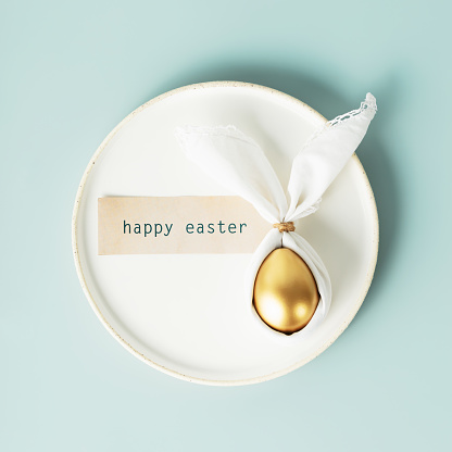 Stylish Easter flat lay with golden egg in easter bunny napkin on white plate over blue background. Minimalist modern Easter table decorations. Top view flat lay