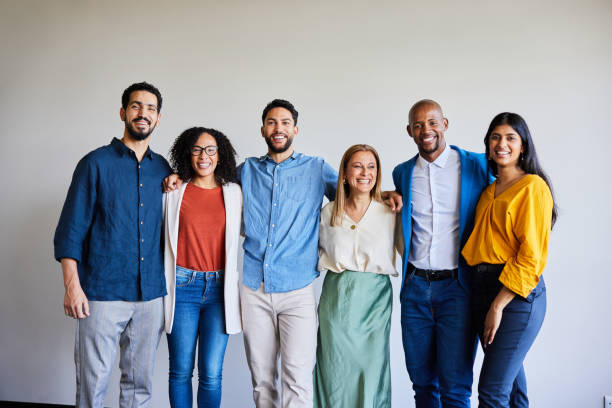 Diverse businesspeople smiling while standing arm in arm in an office Portrait of a smiling group of diverse businesspeople standing arm in arm together in front of an office wall ethnic diversity stock pictures, royalty-free photos & images