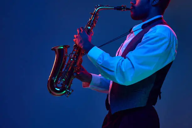 African-American jazz musician playing saxophone his major melodies against blue background in mixed pink-purple lighting. Concept of classical musical instrument, concerts and festivals. Ad