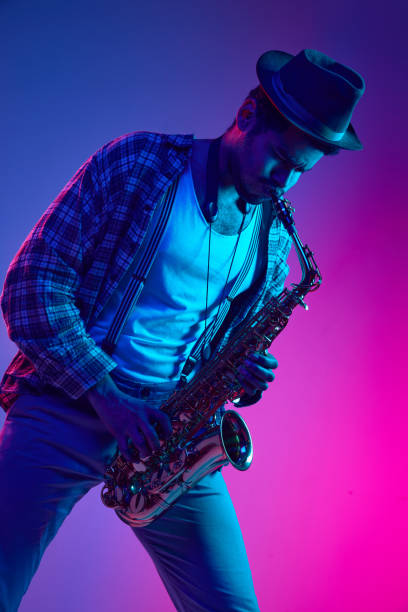 istock African-American man dressed retro, in hat playing saxophone against gradient pink and blue background in neon light. 1977338496