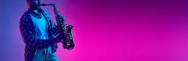 istock Banner. Jazzman playing on saxophone in neon light against gradient blue-pink background with negative space to insert text. 1977338500