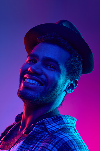 Portrait of African-American artist, Jazzman in hat smiling looking away in neon light against gradient blue-pink background. Concept of jazz, blues, classy instrumental music, festivals and concerts.