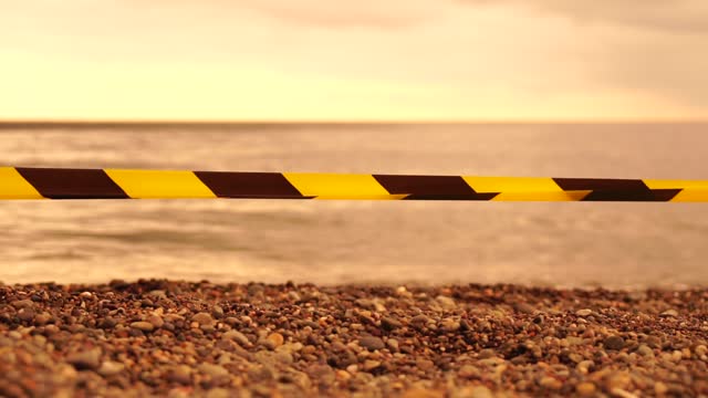 Yellow black warning tape barrier ribbon swinging in the wind across exotic sea beach background without people. No entry Yellow black caution tape. No holiday concept, delayed travel, no summer plans