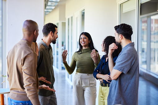 Young businesswoman talking and laughing with a group of diverse coworkers while standing together in the corridor of an office