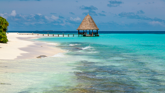 View from shore of sandy beach on wooden jetty over turquoise ocean leading to a thatched pavillon, maldives islands