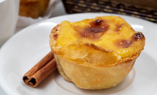 Plate with one Portuguese Pastel de Nata pie with custard and cinnamon sticks. Pastel de Belem is an iconic cup-shaped pastry with a burnt top and a crispy puff pastry crust. Sweet dessert
