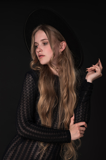 Portrait of shy young blonde looking away, shyly raising one arm bent at elbow, supporting it with other. Blonde with long hair wears short black knitted dress and wide brimmed felt hat.