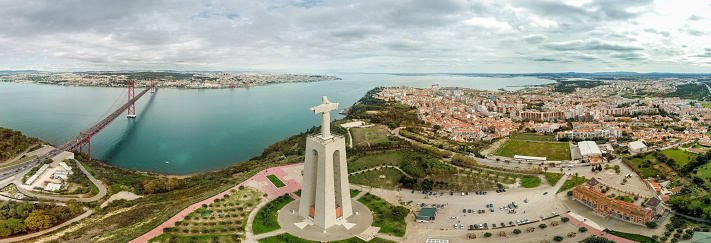 Cinematic aerial perspective of Santuario de Cristo Rei Statue and 25 Abril Bridge. The Statue is situated in Almeda and the bridge connect the Lisbon city with Almada. European travel destination