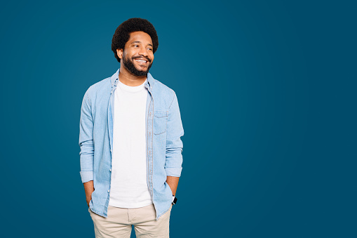 Friendly man with a curly afro hair smiles casually, hands in pockets against a cool blue background, presenting a relaxed yet engaging demeanor. The concept of the casual style of modern casual wear