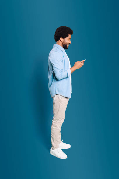 A young man in profile, deeply engaged with his phone, illustrating the ubiquitous nature of mobile devices in modern life A young man in profile, deeply engaged with his phone, illustrating the ubiquitous nature of mobile devices in modern life. Full length photo of Brazilian man using smartphone isolated on blue deep pick stock pictures, royalty-free photos & images