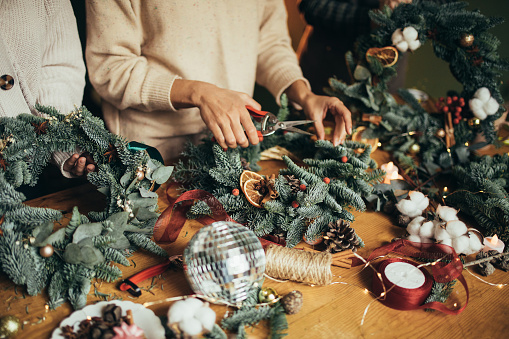 Happy women making Christmas wreath using natural pine branches and festive decorations, standing near wooden table.