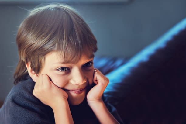 Smiling boy with dreamy look rests his cheeks on his hands while sitting on sofa in a dark room.