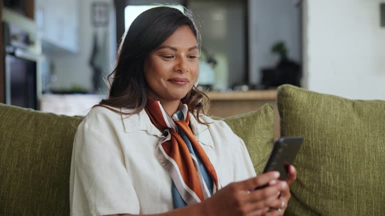 Phone, smile and happy woman on a sofa with social media, chat or web communication in her home. Smartphone, app and Indian female person in a living room with online text, search or gif message